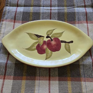 May include: A yellow oval serving platter with a red rim. The platter features a painted design of two red apples with green leaves and a brown stem. The platter is on a plaid tablecloth.