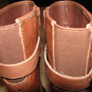 May include: A pair of brown leather boots with a buckle detail. The boots have a brown fabric lining and a zipper on the inside of each boot.