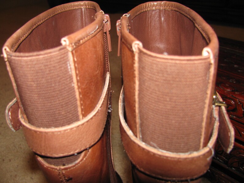 May include: A pair of brown leather boots with a buckle detail. The boots have a brown fabric lining and a zipper on the inside of each boot.