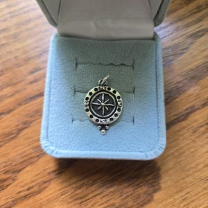 May include: A circular silver pendant featuring a compass rose design with cardinal directions N, E, S, and W engraved around the perimeter. The pendant has a small bail at the top and a decorative three-dot accent at the bottom, resting inside a light blue box.