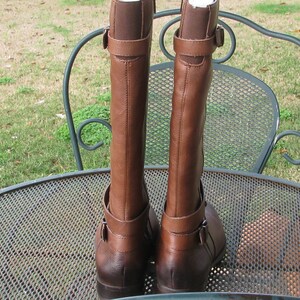 May include: A pair of brown leather riding boots with buckle accents. The boots are on a metal table with a mesh design.