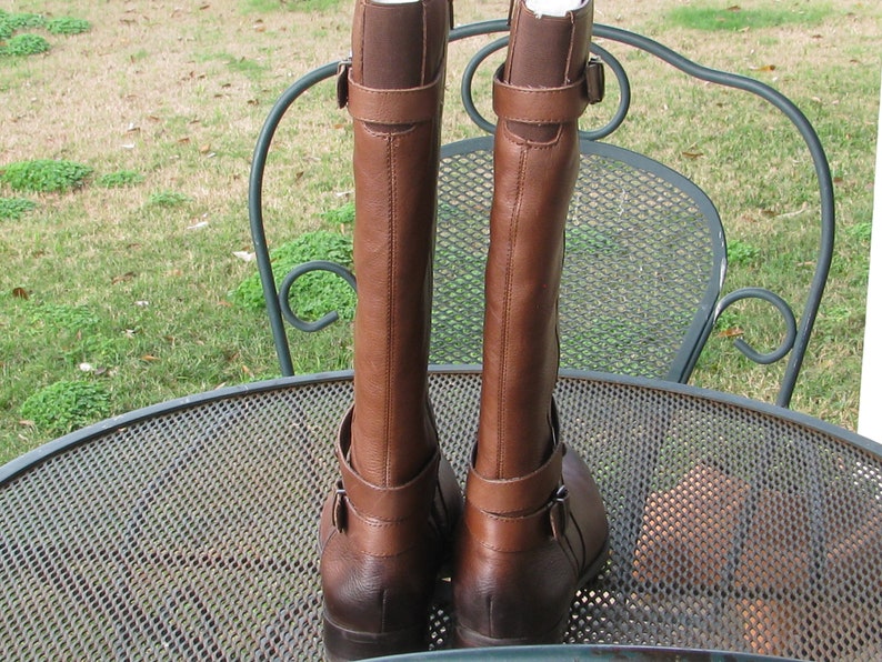 May include: A pair of brown leather riding boots with buckle accents. The boots are on a metal table with a mesh design.
