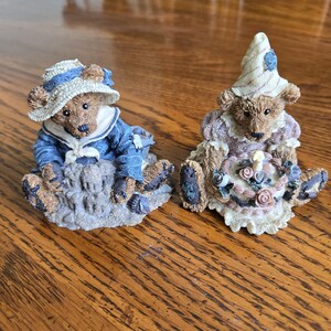 May include: Two decorative bear figurines. One bear wears a blue sailor outfit and hat. The other bear wears a lavender dress and a pointed hat, holding a floral arrangement. Both bears are seated on a textured base.