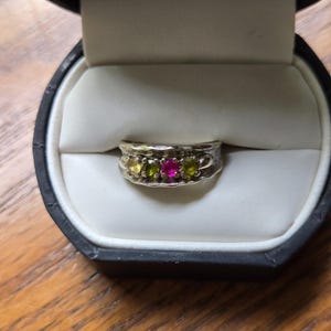 May include: A silver ring with a row of small gemstones in a black ring box. The gemstones are yellow, green, and pink. The ring is set against a white background.