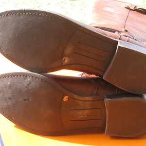 May include: Close-up of the brown leather soles of a pair of brown leather riding boots. The boots have a zipper on the inside of the shaft and a buckle on the outside. The soles are marked "LEATHER CUSHIONING".