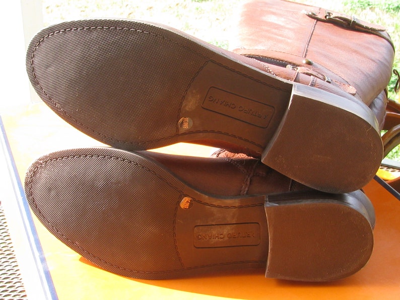 May include: Close-up of the brown leather soles of a pair of brown leather riding boots. The boots have a zipper on the inside of the shaft and a buckle on the outside. The soles are marked "LEATHER CUSHIONING".