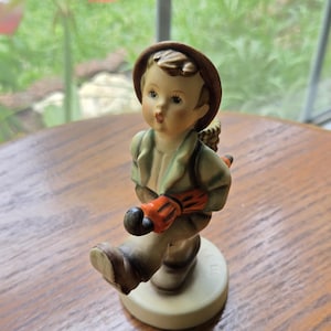 May include: A ceramic figurine of a young person wearing a brown hat and green jacket, holding an orange umbrella. The figurine is on a round, off-white base. The figure is walking with one foot forward.