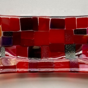 May include: A rectangular glass dish with a mosaic design. The mosaic is made up of small squares of red, orange, purple, and white glass. The dish is slightly curved and has a clear glass base.