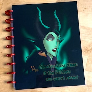 Puede incluir: Una cubierta de agenda negra y verde con una imagen de dibujos animados de Mal&eacute;fica de La Bella Durmiente. La cita "Sometimes the Villain is the Princess who wasn't rescued" est&aacute; impresa en la cubierta.