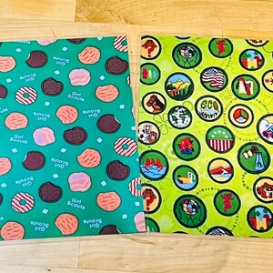 Puede incluir: Dos cuadrados de tela con patrones &uacute;nicos. Uno es verde azulado con ilustraciones de galletas y donuts, y las palabras "Girl Scouts". El otro es verde lima con ilustraciones circulares de varias insignias y la palabra "eco".
