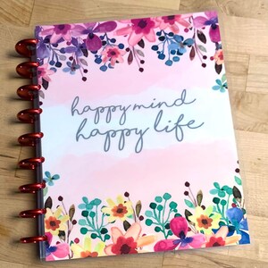 Puede incluir: Un planificador con un dise&ntilde;o floral y las palabras "happy mind happy life" en escritura gris. La portada presenta un dise&ntilde;o estilo acuarela con flores rosas, moradas, azules y amarillas. El planificador tiene anillos met&aacute;licos rojos.