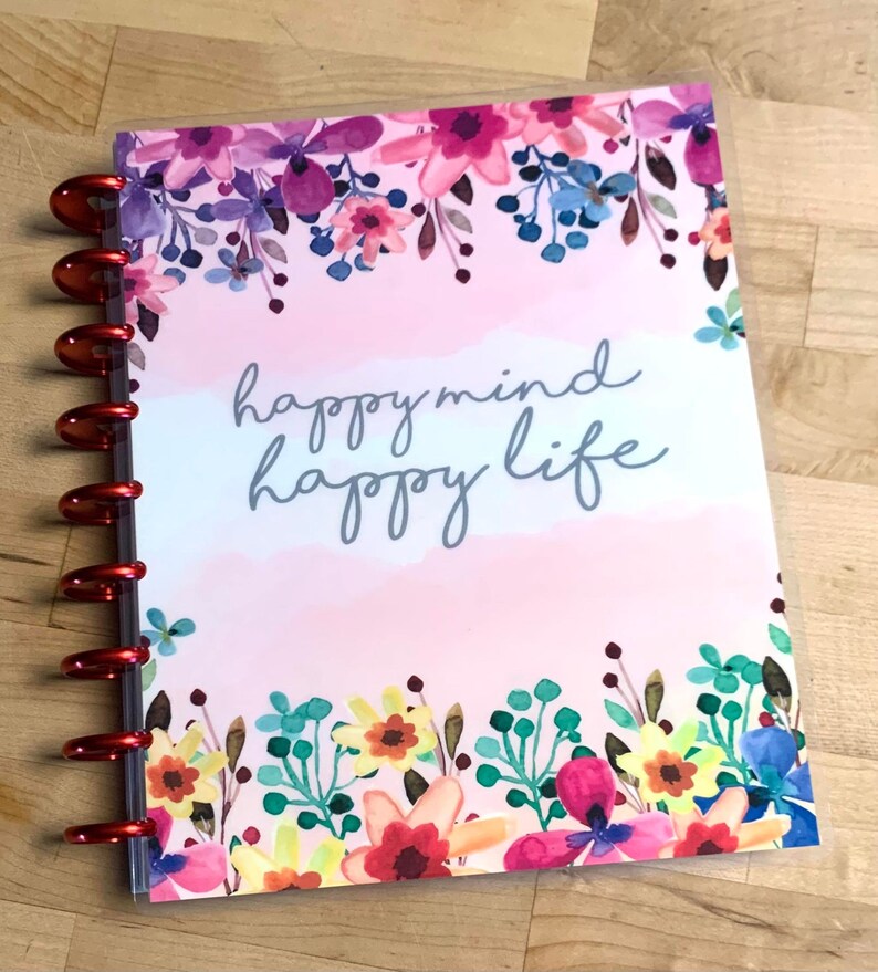 Puede incluir: Un planificador con un dise&ntilde;o floral y las palabras "happy mind happy life" en escritura gris. La portada presenta un dise&ntilde;o estilo acuarela con flores rosas, moradas, azules y amarillas. El planificador tiene anillos met&aacute;licos rojos.