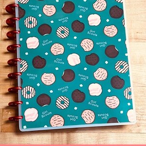 Puede incluir: Un cuaderno turquesa con un estampado de galletas Girl Scout en varios colores. El cuaderno tiene una encuadernaci&oacute;n roja y las palabras "Girl Scouts" est&aacute;n impresas en la portada. La parte inferior del cuaderno tiene las palabras "MATTE FINISH" en texto blanco sobre un fondo rosa.