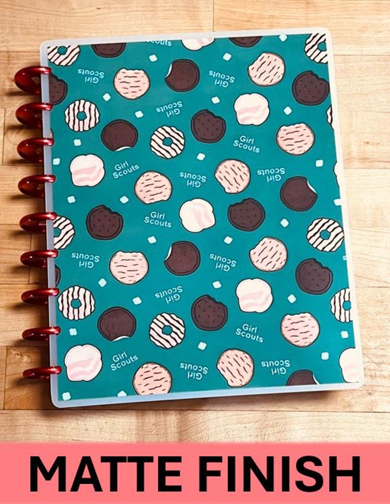 Puede incluir: Un cuaderno turquesa con un estampado de galletas Girl Scout en varios colores. El cuaderno tiene una encuadernaci&oacute;n roja y las palabras "Girl Scouts" est&aacute;n impresas en la portada. La parte inferior del cuaderno tiene las palabras "MATTE FINISH" en texto blanco sobre un fondo rosa.