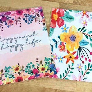Puede incluir: Dos cubiertas de planificador con dise&ntilde;os florales. Una presenta el texto "happymind happy life" sobre un fondo rosa acuarela, enmarcado por flores coloridas. La otra tiene un fondo blanco con un vibrante patr&oacute;n floral.