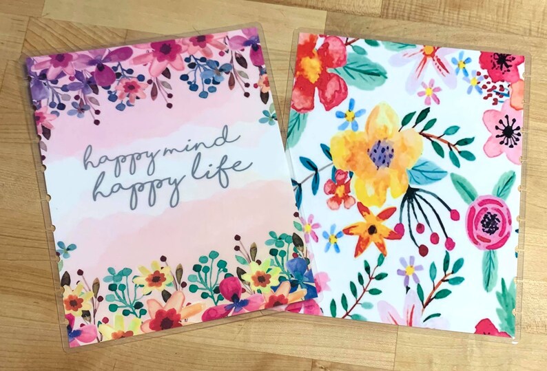 Puede incluir: Dos cubiertas de planificador con dise&ntilde;os florales. Una presenta el texto "happymind happy life" sobre un fondo rosa acuarela, enmarcado por flores coloridas. La otra tiene un fondo blanco con un vibrante patr&oacute;n floral.