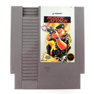 Puede incluir: Cartucho de juego gris para Nintendo Entertainment System (NES) del juego "Rush'n Attack". La etiqueta presenta un soldado apuntando con un arma, con el título del juego en una fuente irregular. El cartucho tiene los logotipos de Nintendo y Konami.