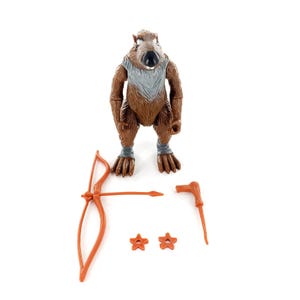May include: A brown anthropomorphic rat action figure with a grey chest patch and grey wrist bands. The figure is accompanied by a set of orange plastic accessories including a bow, a staff, and two small star-shaped throwing weapons.