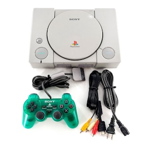 Puede incluir: Una consola de videojuegos Sony PlayStation de color gris claro con una tapa circular para discos, un mando con cable de color verde translúcido, un cable AV gris y un cable de alimentación negro. La consola incluye el logotipo de Sony, el logotipo de PlayStation y botones etiquetados como Reset y Open.