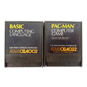 May include: Two black Atari game cartridges. The left cartridge is labeled BASIC COMPUTING LANGUAGE, USE WITH CONSOLE KEYBOARD, ATARI CXL4002. The right cartridge is labeled PAC-MAN COMPUTER GAME, USE WITH JOYSTICK CONTROLLERS, ATARI CXL4022.