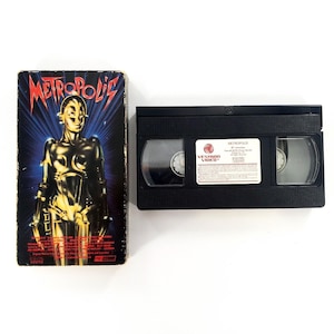 Może przedstawiać: Winylowa kaseta VHS z filmu "Metropolis" z oryginalnym pudełkiem. Pudełko przedstawia stylizowany obraz złotego robota na niebiesko-czerwonym tle. Czarna kaseta VHS znajduje się obok pudełka.