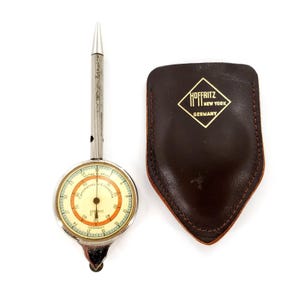 May include: A silver-toned map measurer with a circular dial featuring scales for inches, miles, centimeters, and kilometers. The device has a long handle and a small wheel at the base. Beside it is a dark brown leather case with a gold logo reading Hoffritz New York Germany.