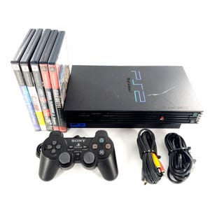 Op de afbeelding: Een zwarte PlayStation 2 console met een bijpassende zwarte Sony controller, twee zwarte stroom- en AV-kabels, en vijf videospelhoesjes waaronder Dance Dance Revolution Extreme 2, NBA Live 06, Ford Mustang, Gran Turismo 3 en Call of Duty 3.