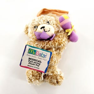 May include: A tan plush teddy bear toy with a purple fabric collar and a purple, yellow, and pink rattle accessory. A tag attached to the bear reads McBaby Always Quality. Always Fun. MUSICAL Pull-string Plush Toy (Birth to 18 mos).
