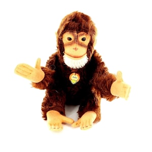 Puede incluir: Un peluche de orangután marrón con cara marrón claro, manos y pies beige y cuello blanco. El juguete tiene una etiqueta que dice "Jocko". El orangután está sentado con los brazos extendidos.