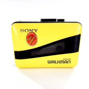 May include: A yellow Sony Walkman cassette player with black accents. The device has the word "SONY" printed on the top and "WALKMAN" on the bottom. A red and gold sticker reads "Digital Synthesizer aiwa".