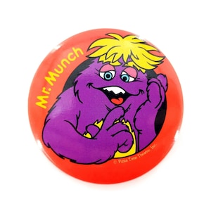 May include: A round, red button featuring a cartoon character named Mr. Munch. The character is purple with yellow hair, blue eyes, and a smiling expression. The button has the text "Mr. Munch" and "Pizza Time Theatre Inc."