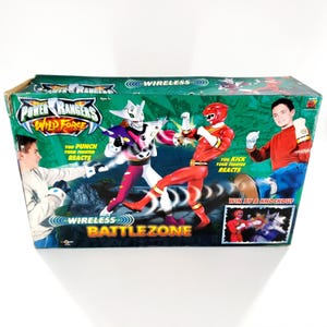 May include: A Power Rangers Wild Force Wireless Battlezone toy box. The box features images of Power Rangers characters in action, with text that reads "You Punch Your Fighter Reacts" and "You Kick Your Fighter Reacts". The box is green and blue.