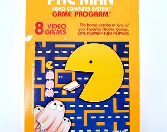 Puede incluir: Una caja de cartucho de videojuego Pac-Man vintage. La caja es naranja con el logotipo de Pac-Man y detalles del juego. El juego presenta un personaje Pac-Man amarillo, fantasmas y un diseño de laberinto. La caja dice "8 Videojuegos" y "Programa de juego del sistema informático de video".