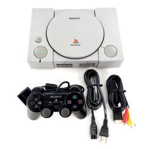 May include: A light gray Sony PlayStation game console with a circular disc lid, featuring the Sony logo and PlayStation branding. Included is a black wired controller with dual analog sticks and a set of black power and AV cables with yellow and red plugs.