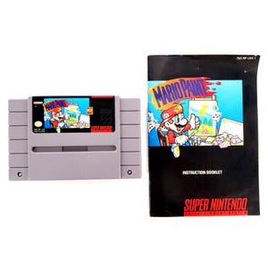以下が含まれることがあります： マリオペイントのグレーのスーパーファミコン用ゲームカセットと、黒い取扱説明書のセット。カセットのラベルと説明書の表紙には、イーゼルの前で絵を描くマリオのカラフルなイラストと「MARIO PAINT」の文字が印刷されています。