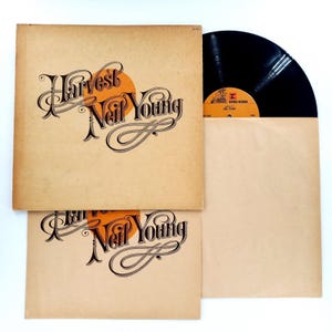May include: A Neil Young Harvest vinyl record set featuring a tan cardboard album cover with black cursive text and an orange circle graphic. The set includes a black vinyl disc with an orange Reprise Records label and a matching tan inner sleeve.