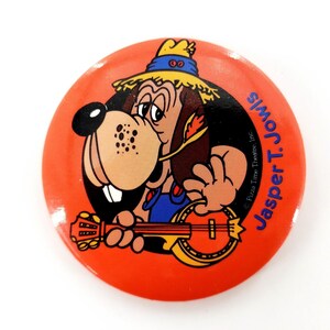 May include: A round, orange button featuring a cartoon dog playing a banjo. The dog wears a straw hat and overalls. Text on the button reads "Jasper T. Jowls" and "Pizza Time Theatre, Inc."