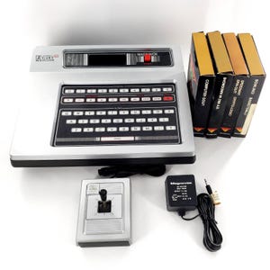 May include: A silver Magnavox Odyssey2 video game console with a QWERTY keyboard, a single joystick controller, a black power adapter, and four game cartridges labeled Computer Golf, Showdown in 2100 A.D., Speedway, and Bowling/Basketball.