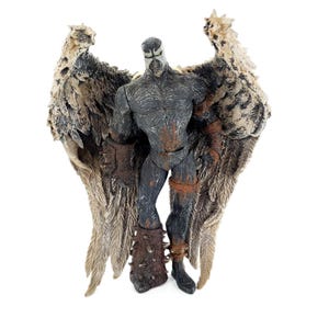 May include: A detailed action figure of a dark-skinned character with large, feathered wings. The figure has a textured, muscular physique and is adorned with brown and rust-colored accents. The wings are a mix of brown, tan, and cream colors.