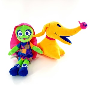Può includere: Due peluche: una bambola dalla pelle verde con i capelli rosa e un cane giallo. La bambola ha tratti cuciti e un vestito patchwork. Il cane ha un lungo muso e un collare rosa, con una piccola zucca sul naso.