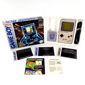 May include: A vintage Nintendo Game Boy Compact Video Game System, complete with the original box, game cartridge, instruction booklets, and a black cable. The box features the Game Boy logo and a graphic of the device. The Game Boy itself is a light gray color.