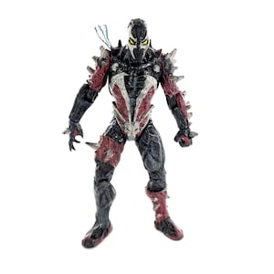 Peut inclure: Figurine d'action détaillée de Spawn, un personnage de bande dessinée, dans une pose dynamique. La figurine est principalement noire et rouge, avec des accents argentés et une armure à pointes. Elle présente un masque avec des yeux jaunes brillants et un effet d'énergie bleue.