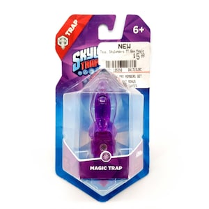 May include: A purple "Magic Trap" toy from the Skylanders series, in a clear plastic case with a purple and blue backing. The packaging features the "Skylanders Trap" logo and the text "6+".