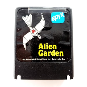 Könnte beinhalten: Schwarze Videospielkassette mit dem Titel "Alien Garden" in gelber Schrift. Die Kassette zeigt ein weißes geometrisches Vogeldesign mit einem roten Herzen. Das Epyx-Logo befindet sich in der oberen rechten Ecke.
