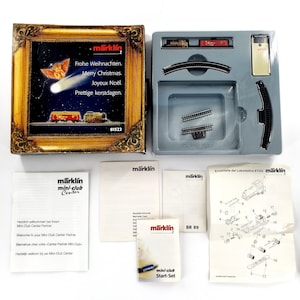 Puede incluir: Un juego de trenes modelo Märklin Z, que incluye una caja con una ilustración festiva y texto en varios idiomas. El juego incluye vías, un vagón de tren y una unidad de control. También se incluyen varios manuales de instrucciones.