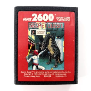 May include: A black Atari 2600 video game cartridge for Secret Quest. The red label features a character with a red helmet holding a yellow staff facing a large dark creature. Text includes Atari 2600, Secret Quest, and copyright information.