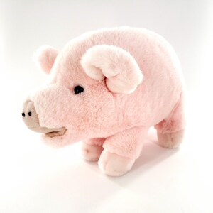 May include: A plush pink pig toy with a soft, fuzzy texture. The pig has small black eyes, a light-colored snout, and small ears. The toy is standing on four short legs.