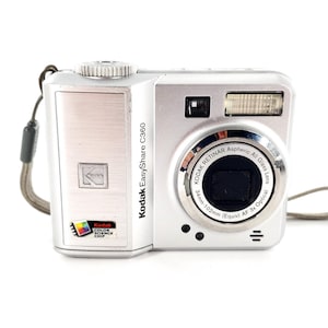 May include: A silver Kodak EasyShare C360 digital camera with a wrist strap. The front features a Kodak logo, a Color Science Chip label, and a lens labeled Kodak Retinar Aspheric All Glass Lens 34mm-102mm (Equiv) AF 3x Optical.