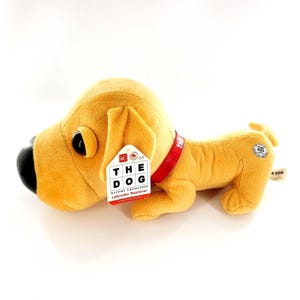 Puede incluir: Un peluche de perro Labrador Retriever dorado con una nariz negra y un collar rojo. El perro tiene una etiqueta que dice "THE DOG Artist Collection Labrador Retriever". El juguete está tumbado sobre una superficie blanca.