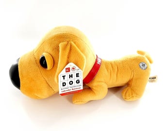 May include: A plush golden Labrador Retriever dog toy with a black nose and a red collar. The dog has a tag that reads "THE DOG Artist Collection Labrador Retriever". The toy is lying down on a white surface.